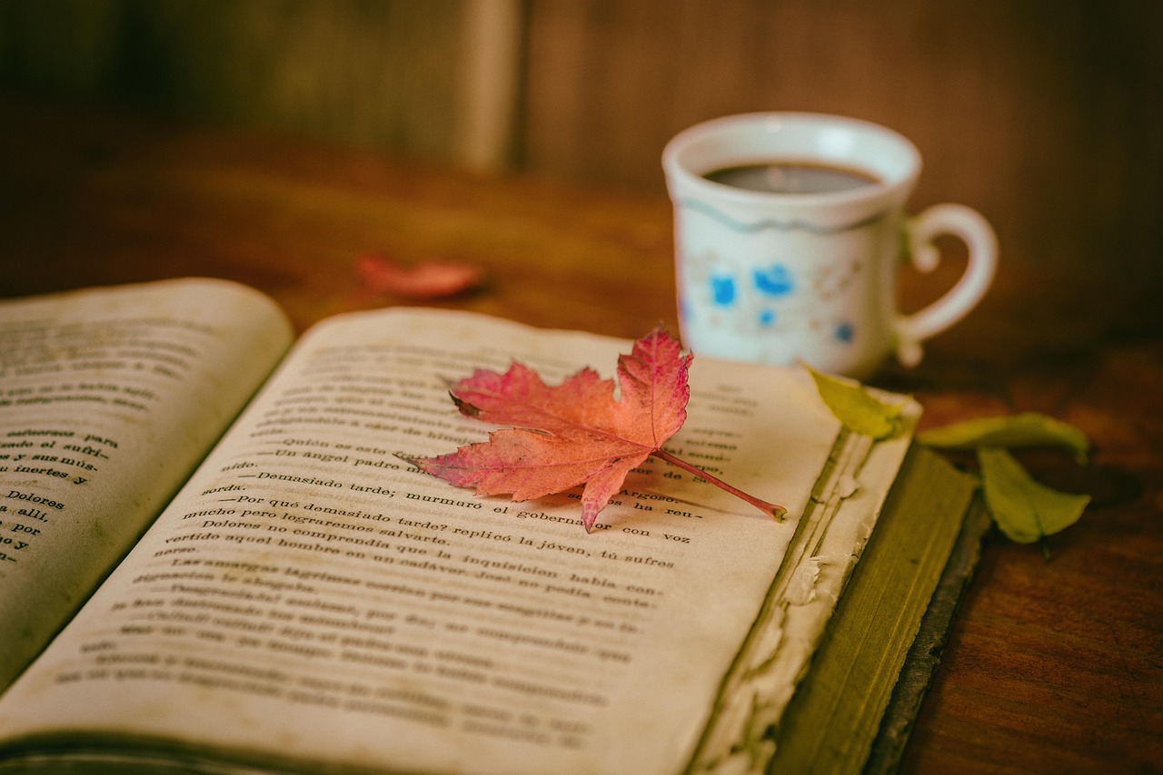 Foto zeigt ein aufgeschlagenes Buch, auf dem ein Ahornblatt liegt. Daneben steht eine Tasse mit Kaffee.
